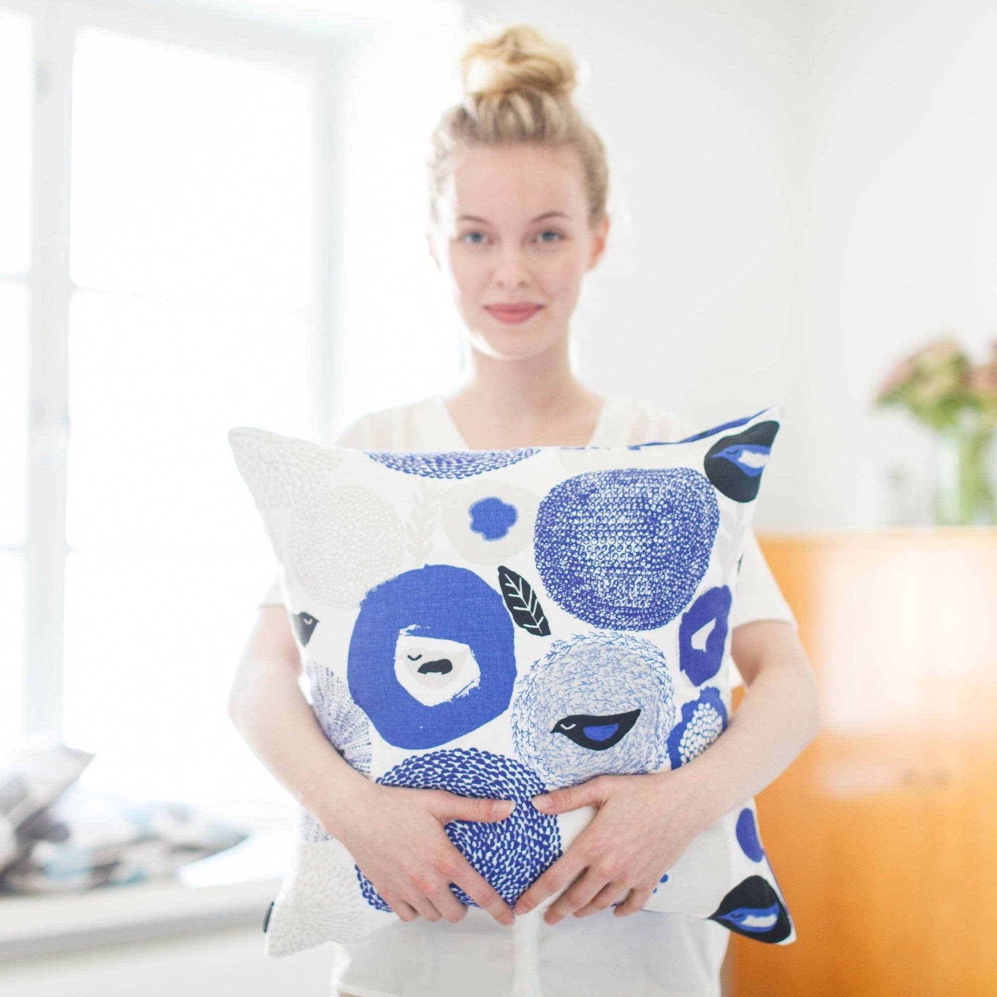 Woman holding blue floral and bird-pattern linen-cotton cushion cover by Matti Pikkujämsä, 18.5" square.