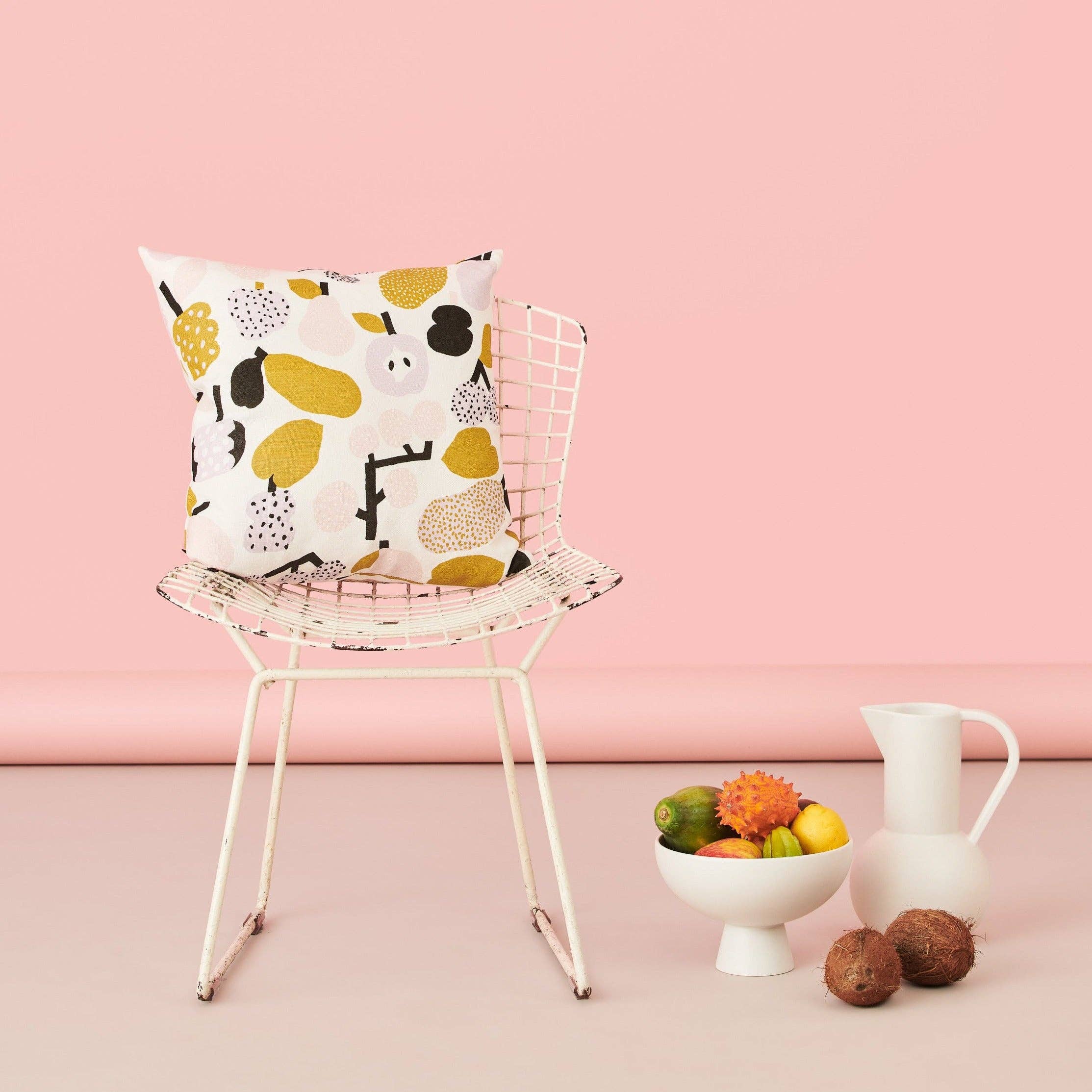 Olive green fruit-pattern linen-cotton cushion cover by Hannele Äijälä, 18.5" square on a wire chair against a pink background.