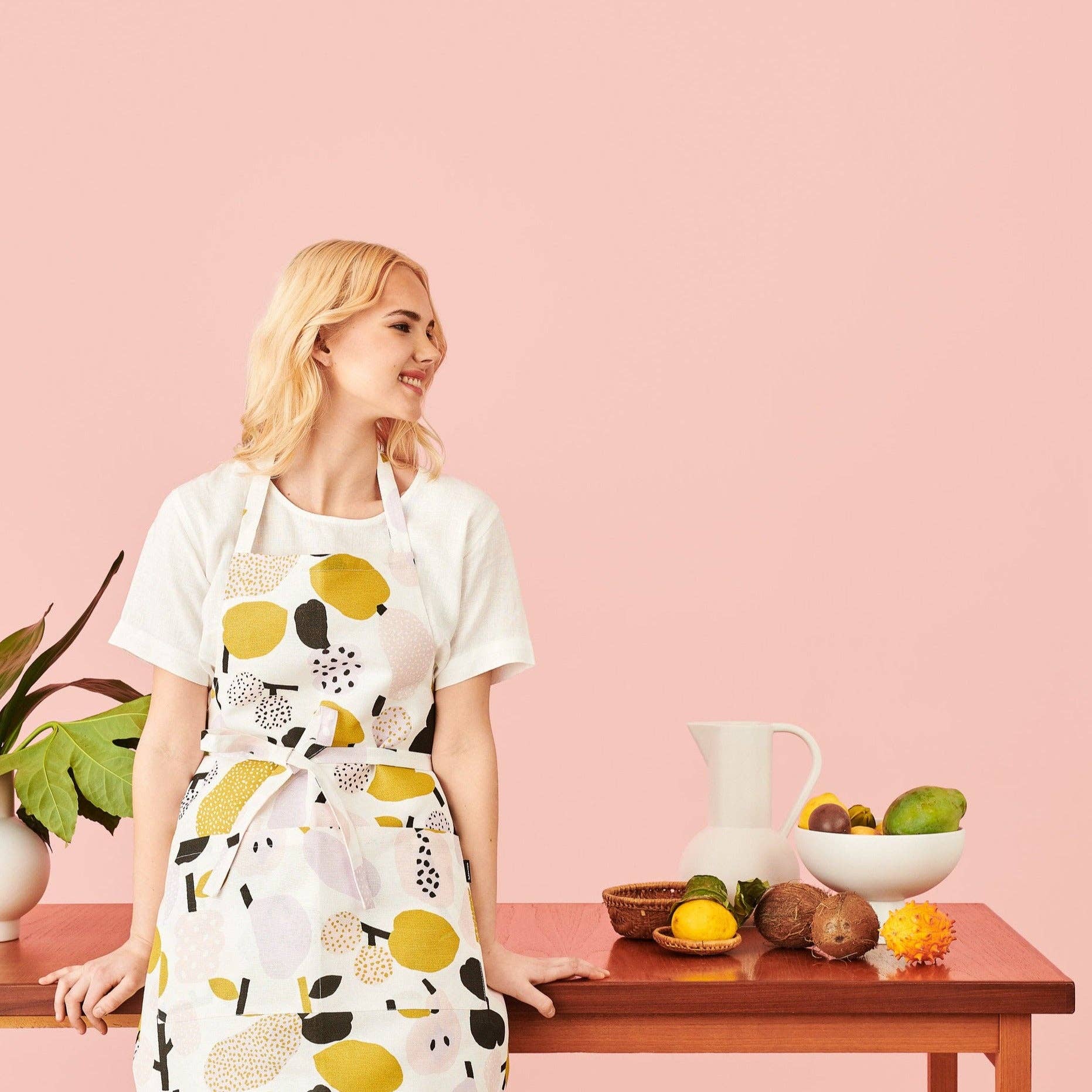 Woman wearing an olive green fruit-pattern linen-cotton apron by Hannele Äijälä standing next to a table with fruits and a pink background.