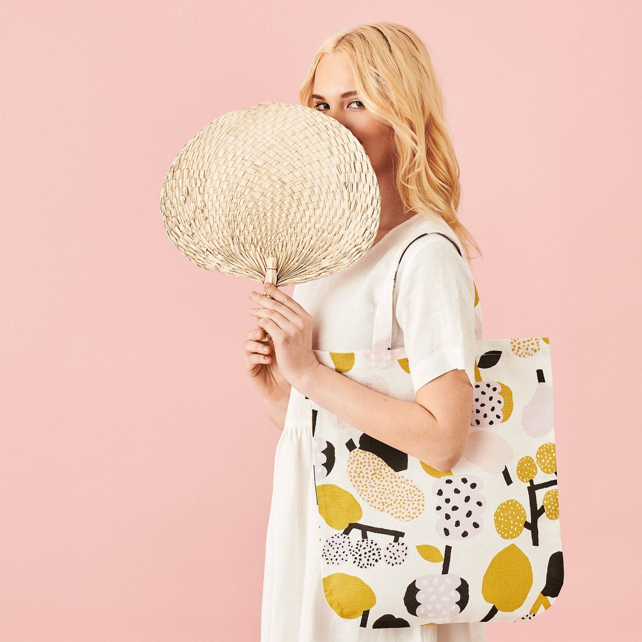 Woman holding an olive green fruit-pattern linen-cotton tote bag by Hannele Äijälä, 15" x 17" against a pink background.