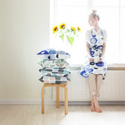 A woman with a stack of patterned pillows and wearing a blue floral and bird-pattern linen-cotton apron by Matti Pikkujämsä..