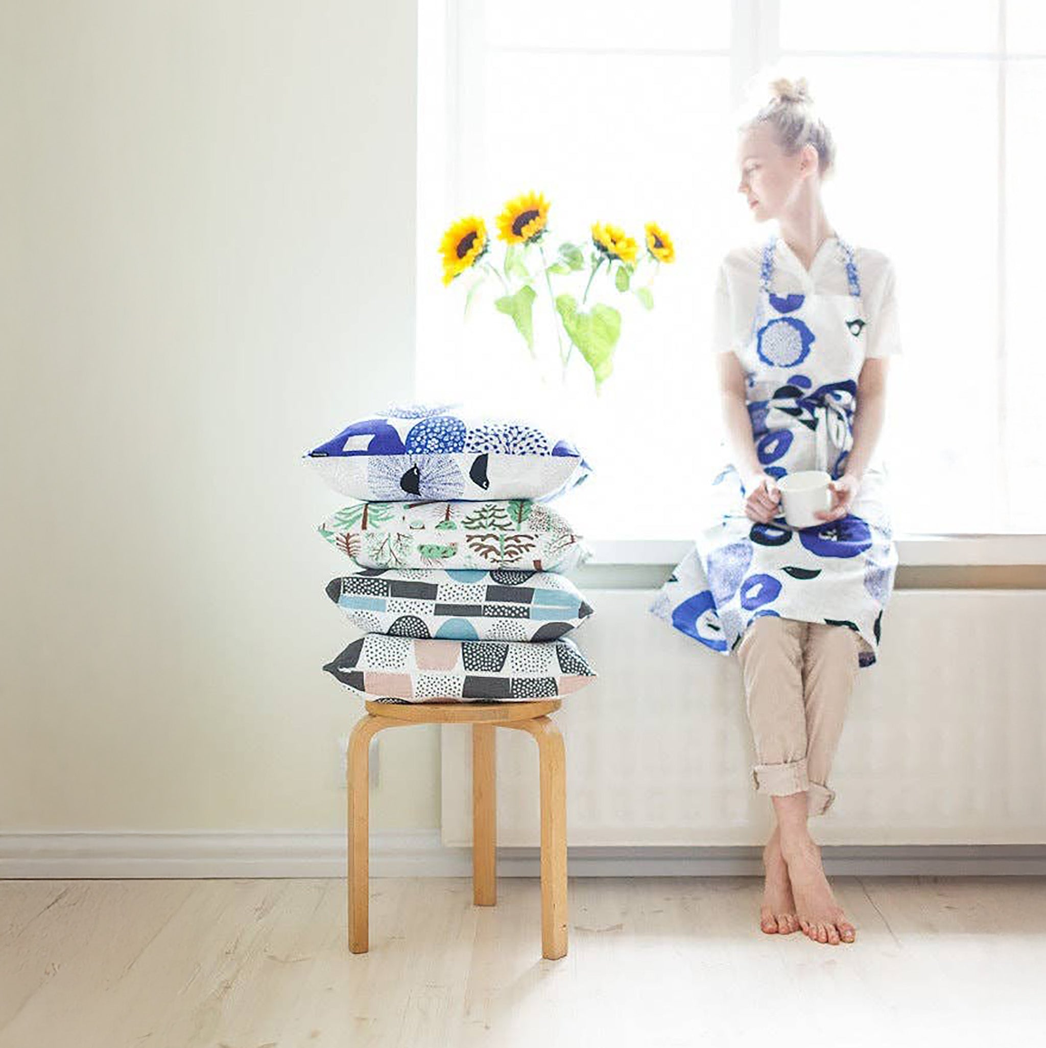 A woman with a stack of patterned pillows and wearing a blue floral and bird-pattern linen-cotton apron by Matti Pikkujämsä..